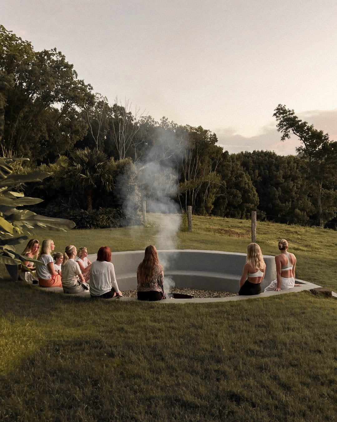 Group of people sitting around a fire pit in a natural setting with trees and grass.
