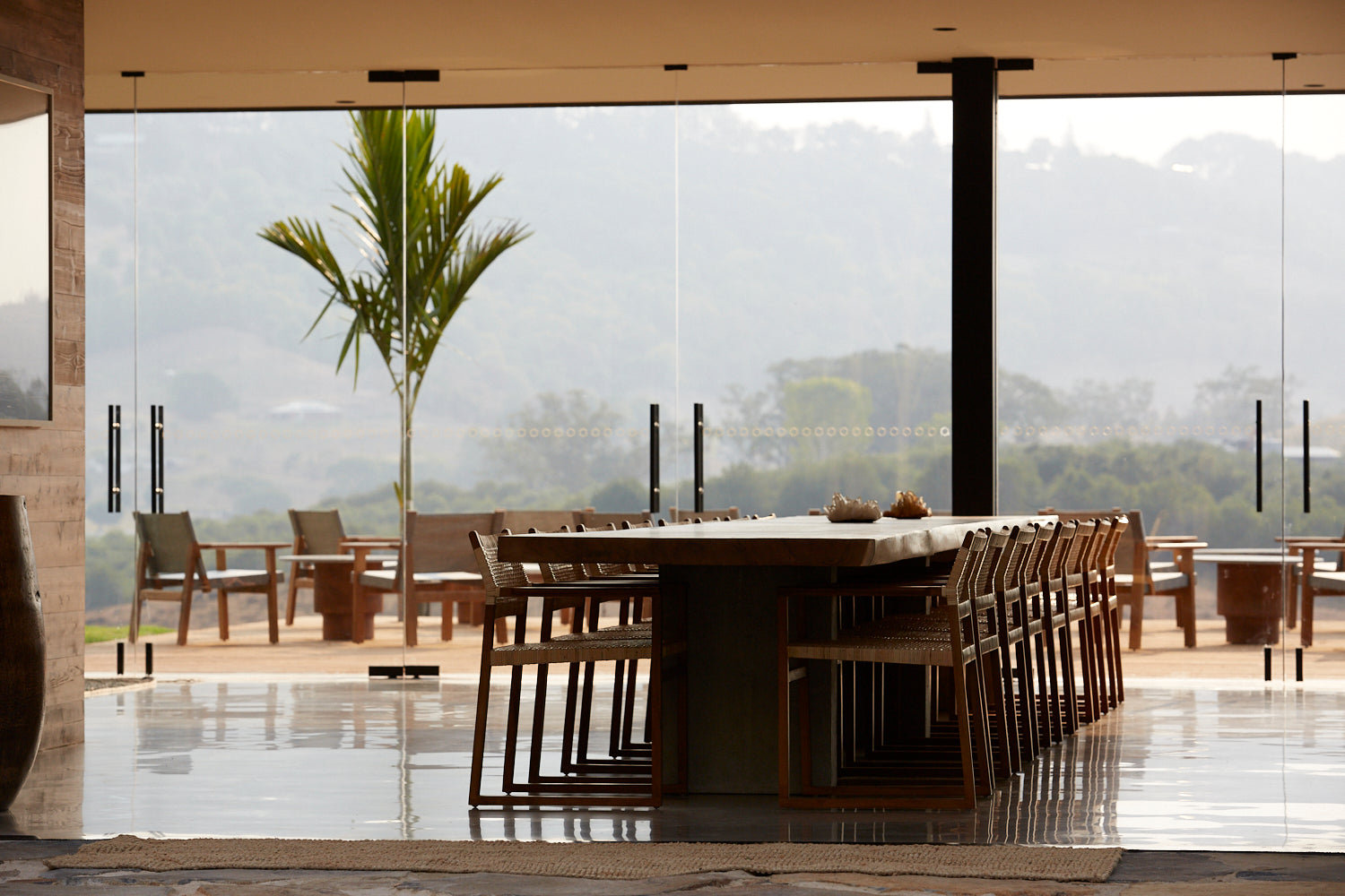 A stone-topped communal table with timber armchairs set back from a wall of frameless glass, slim black pendant cylinders overhead, a young palm rising through the terrace and hazy hills beyond.