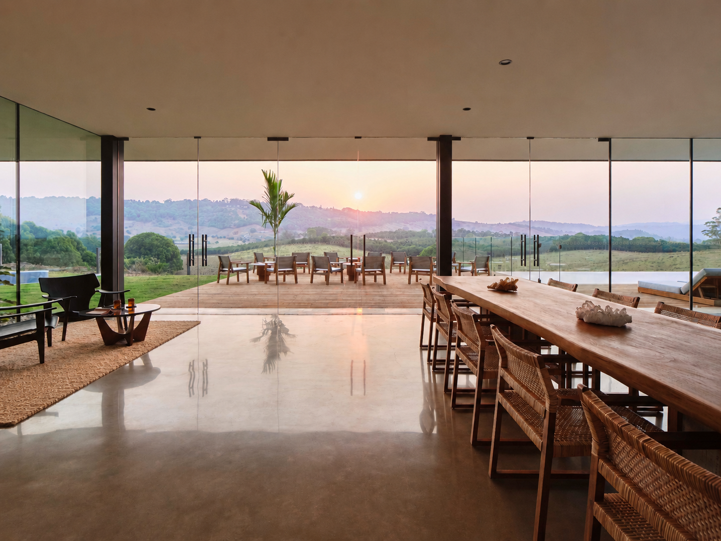 The main house dining room at sunset, a long solid-timber table with rattan chairs on polished concrete, glass walls slid back onto a terrace of dining chairs and the sun dropping behind the hinterland ridges.