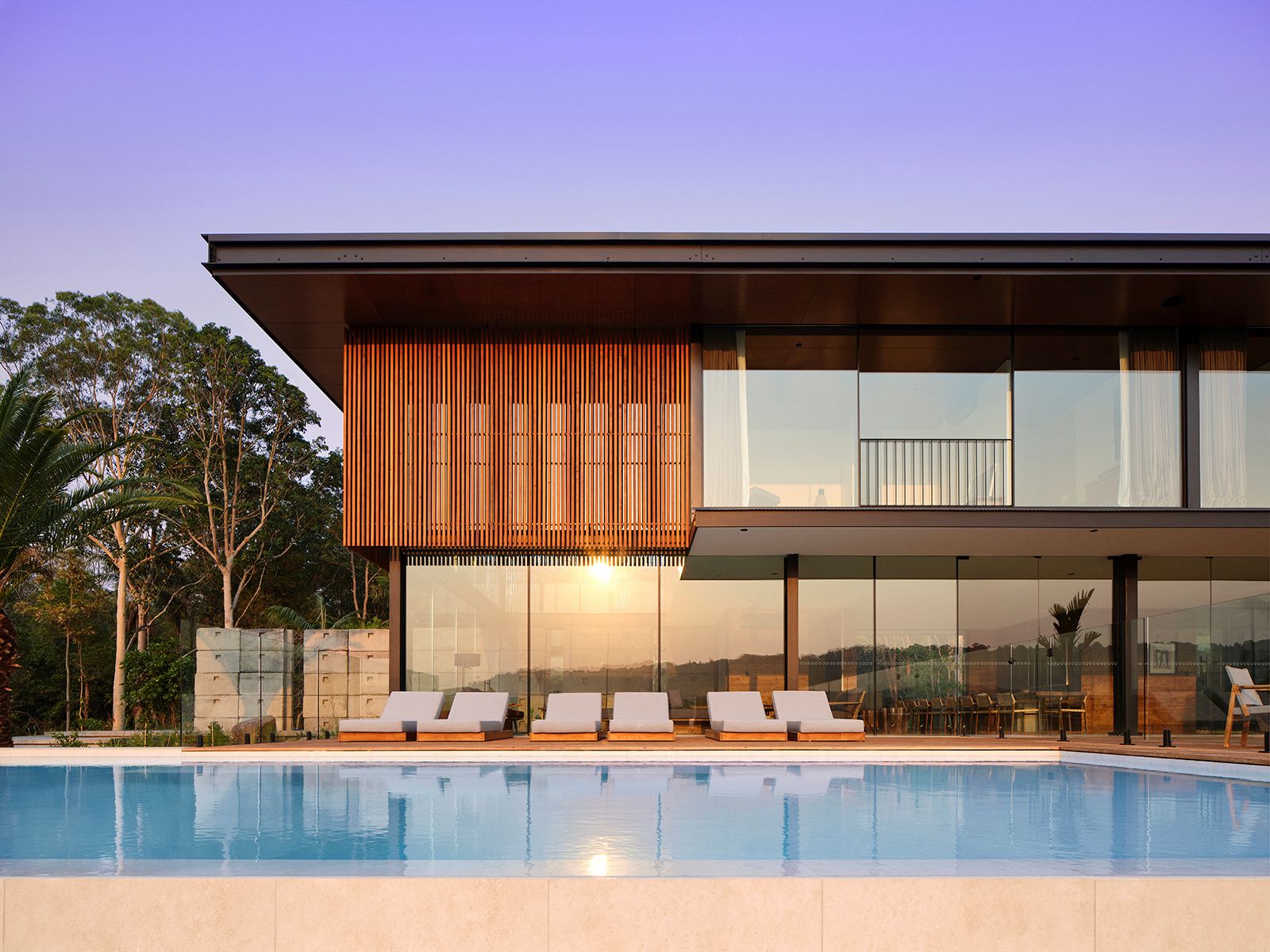 The pool under a violet dusk sky, its still water reflecting a house of ribbed timber battens and glass with white sun lounges lined along the stone coping and the last light burning through the ground-floor glazing.