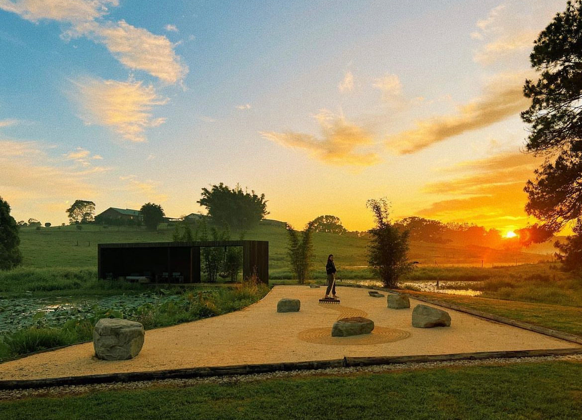 The Japanese garden at sunset, raked gravel swirling between placed boulders, a low dark steel pavilion open to the lily pond on one side and the dam and pasture running out to an orange horizon.