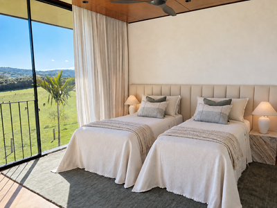 A bright, modern bedroom featuring two neatly made twin beds with cream bedding and gray accent pillows, positioned beneath a beige tufted headboard. The room has a large floor-to-ceiling window looking out onto a lush green landscape, with cream curtains, bedside tables with lamps, and a gray rug.
