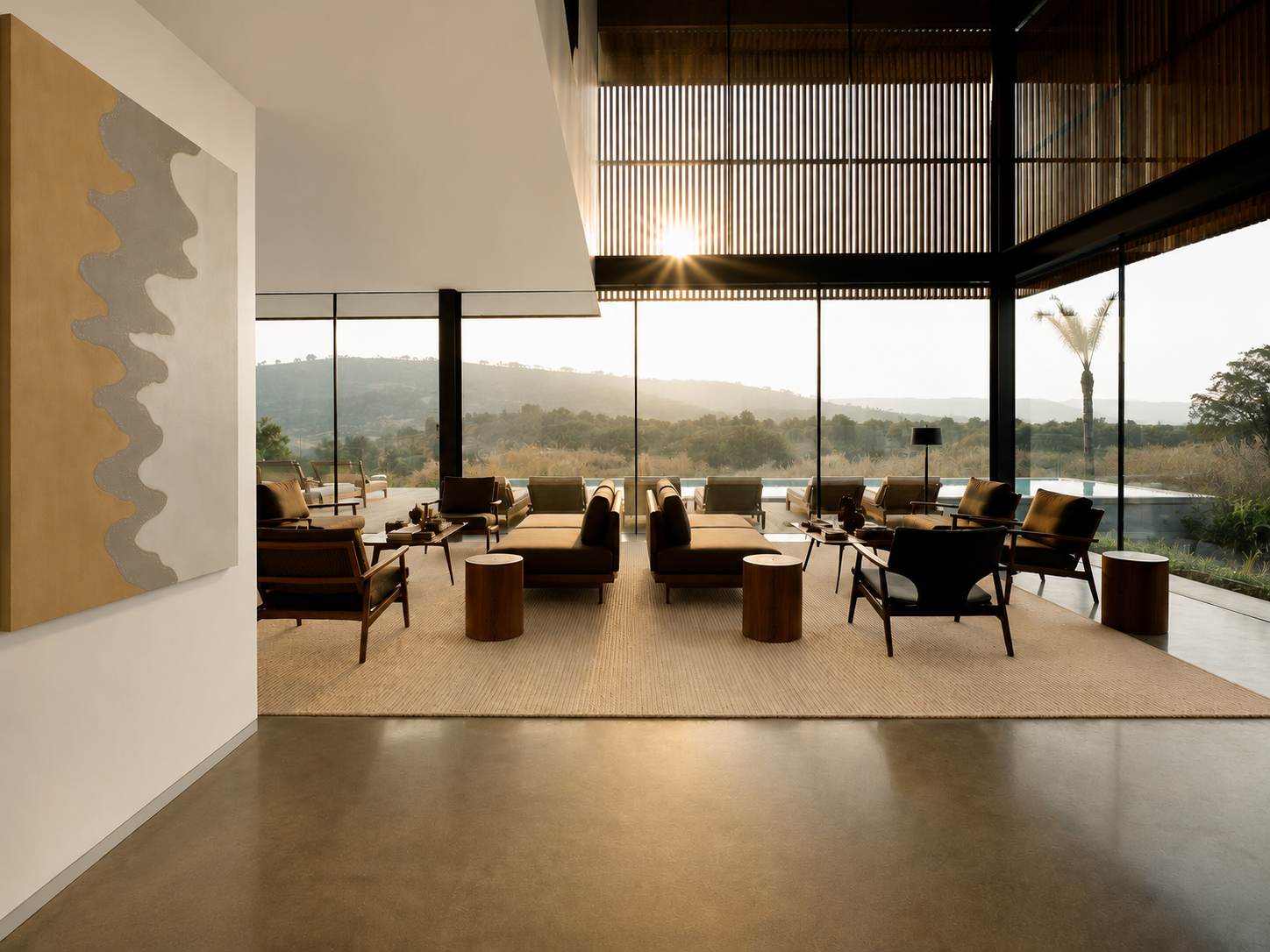 The double-height living room, polished concrete underfoot and a timber-slatted mezzanine screen above, low tan daybeds and cane-backed armchairs on a jute rug facing glass walls that open onto the pool and the hills at sunset.