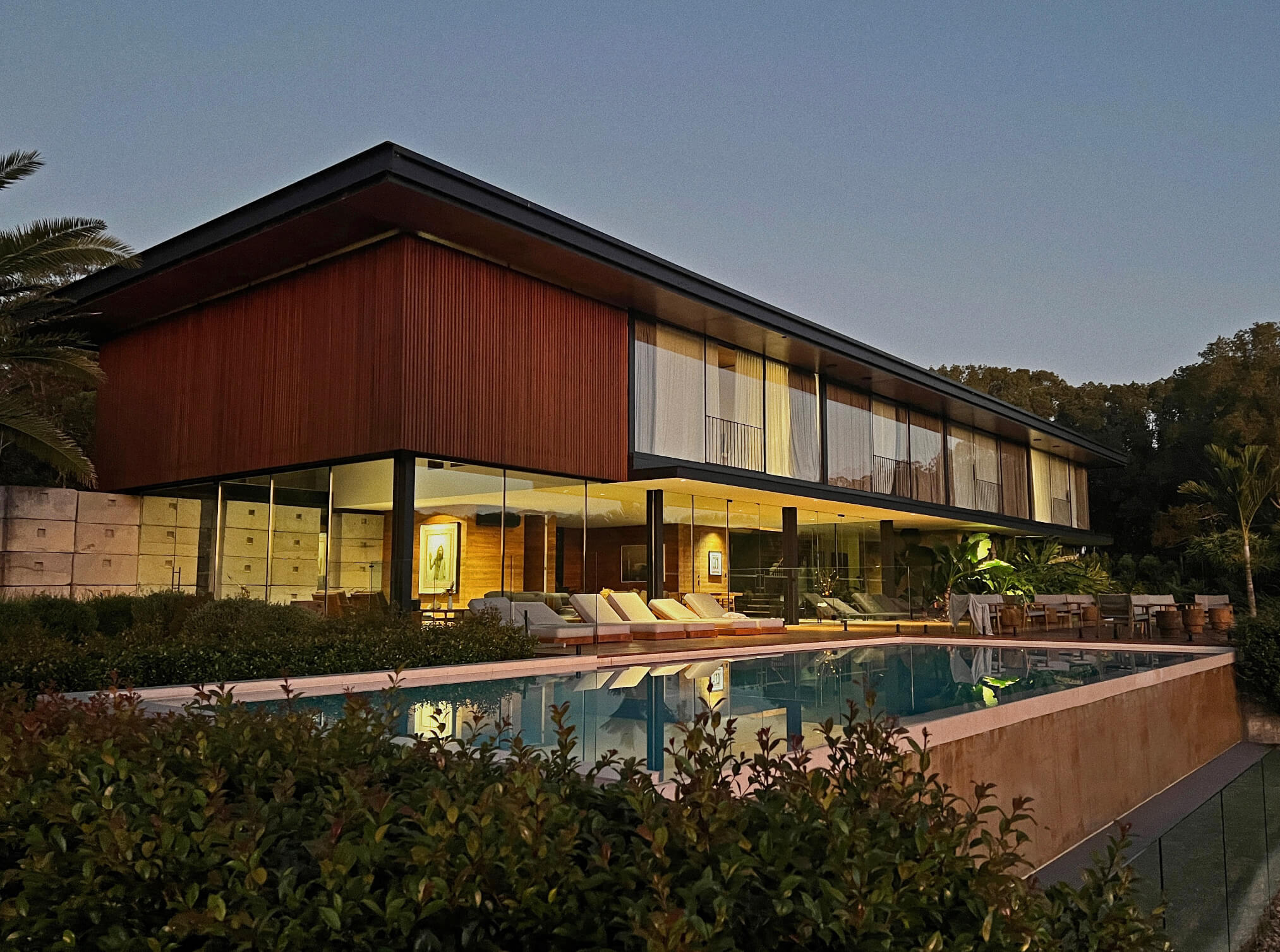 The residence at dusk seen across the lap pool, its long ribbed-timber upper storey cantilevered over a glass ground floor lit warm from within, sun lounges and dining settings ranged along the terrace behind a clipped hedge.
